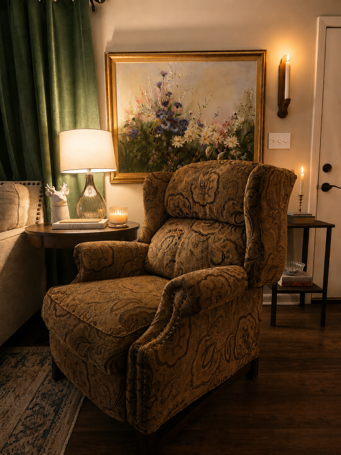 Cozy paisley wingback chair by candlelight with a large floral oil painting, green velvet curtains, and a glass lamp
