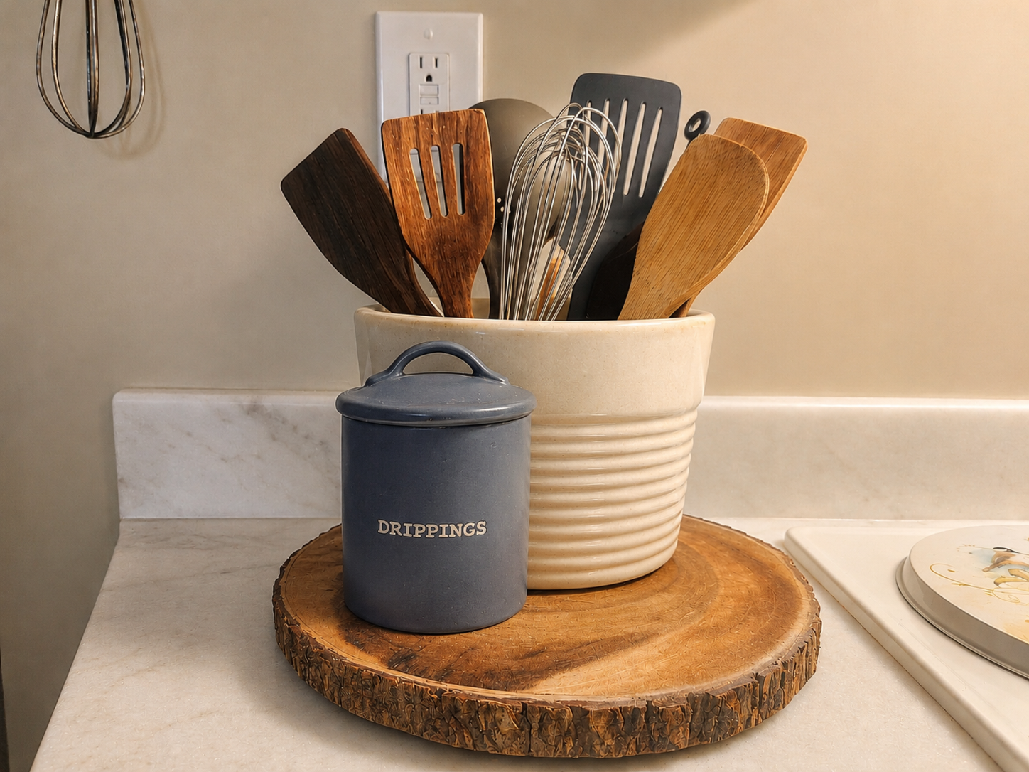 Kitchen counter with a cream ribbed utensil crock full of wooden spoons and spatulas beside a blue drippings jar on a live edge wood board