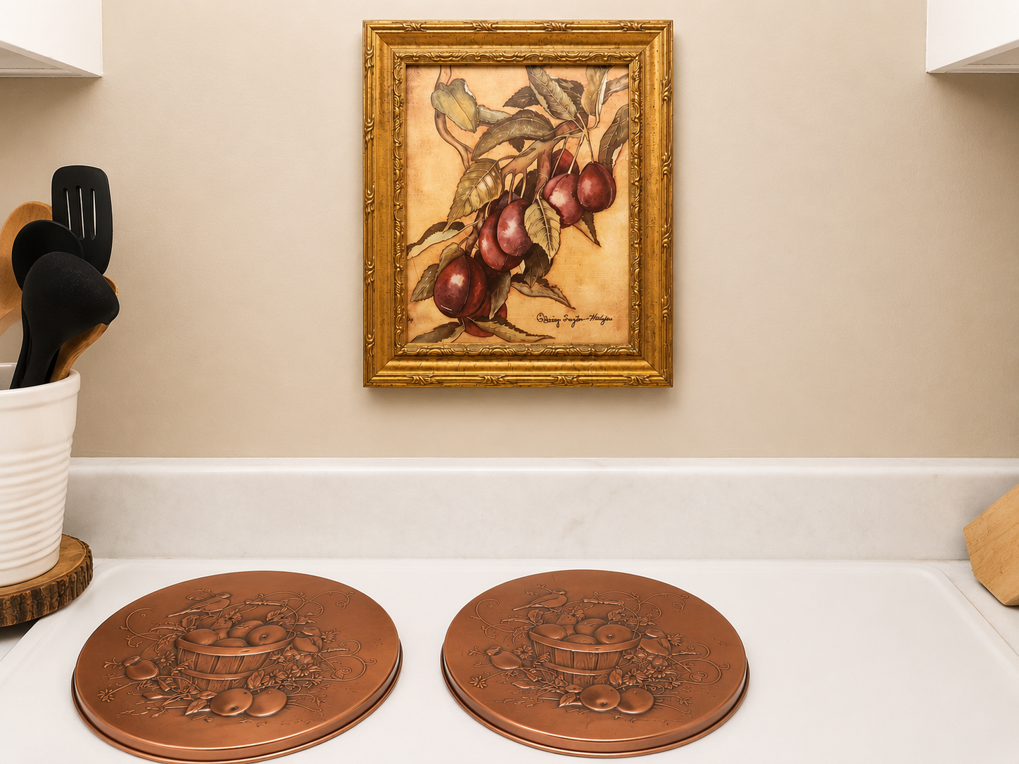Kitchen stove area with a gold-framed fruit painting above copper decorative burner covers and wooden utensils